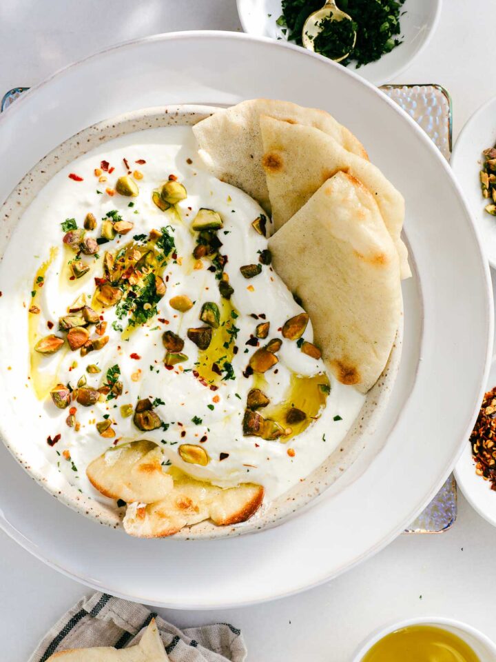 A shallow white bowl filled with thick and creamy lemony whipped feta topped with chopped pistachios, red pepper flakes, and minced parsley.
