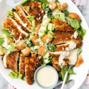Caesar Salad with Crispy Chicken arranged on a white platter with romaine lettuce tossed with caesar dressing and sliced crispy chicken breast.
