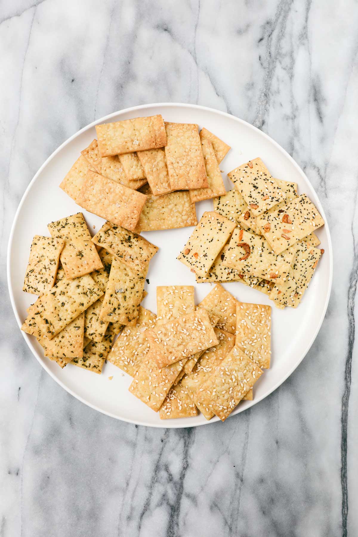 A plate with Einkorn crackers with four different toppings, everything bagel seasoning, flaky sea salt, sesame seeds, and rosemary.