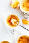 Several ramekins with orange creme brulee with a torched caramelized top broken by a spoon holding a scoop of creme brulee with decorative orange peels and dappled sunlight.