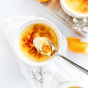 Several ramekins with orange creme brulee with a torched caramelized top broken by a spoon holding a scoop of creme brulee with decorative orange peels and dappled sunlight.