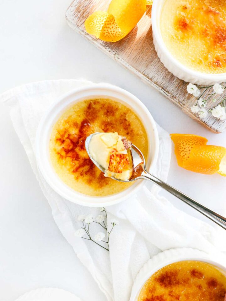 Several ramekins with orange creme brulee with a torched caramelized top broken by a spoon holding a scoop of creme brulee with decorative orange peels and dappled sunlight.