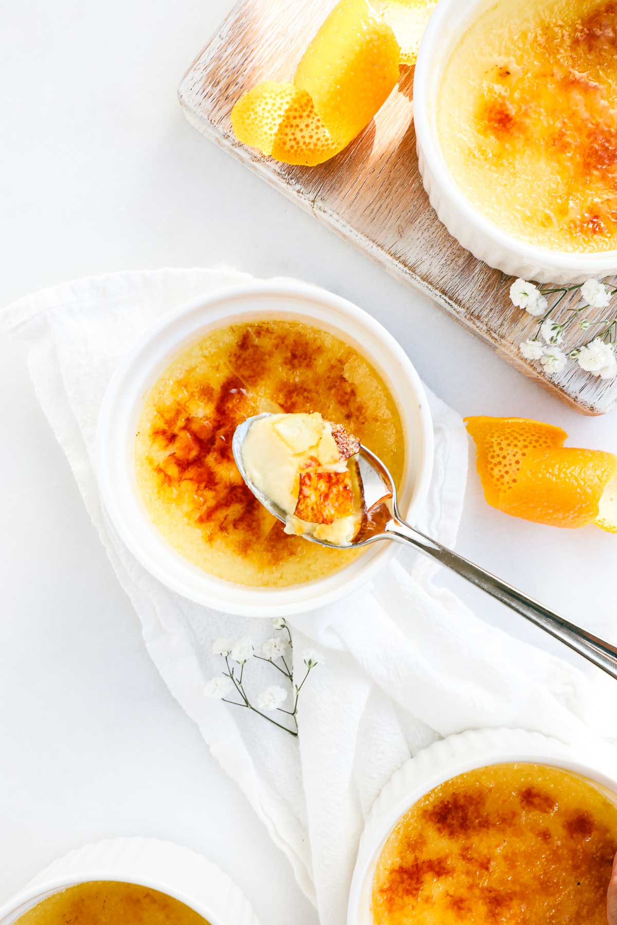 Several ramekins with orange creme brulee with a torched caramelized top broken by a spoon holding a scoop of creme brulee with decorative orange peels and dappled sunlight.