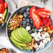 Strawberry and Blueberry salad in a blue bowl featuring candied almonds, sliced avocado, strawberries, blueberries, and goat chees on top of mixed greens.