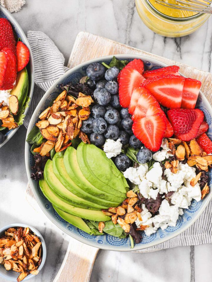 Strawberry and Blueberry salad in a blue bowl featuring candied almonds, sliced avocado, strawberries, blueberries, and goat chees on top of mixed greens.
