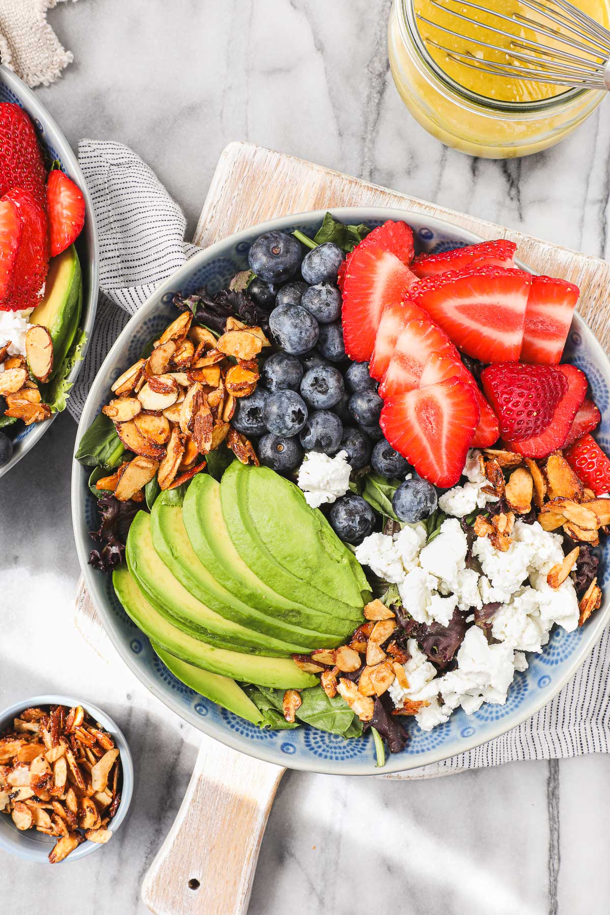 Strawberry and Blueberry salad in a blue bowl featuring candied almonds, sliced avocado, strawberries, blueberries, and goat chees on top of mixed greens.