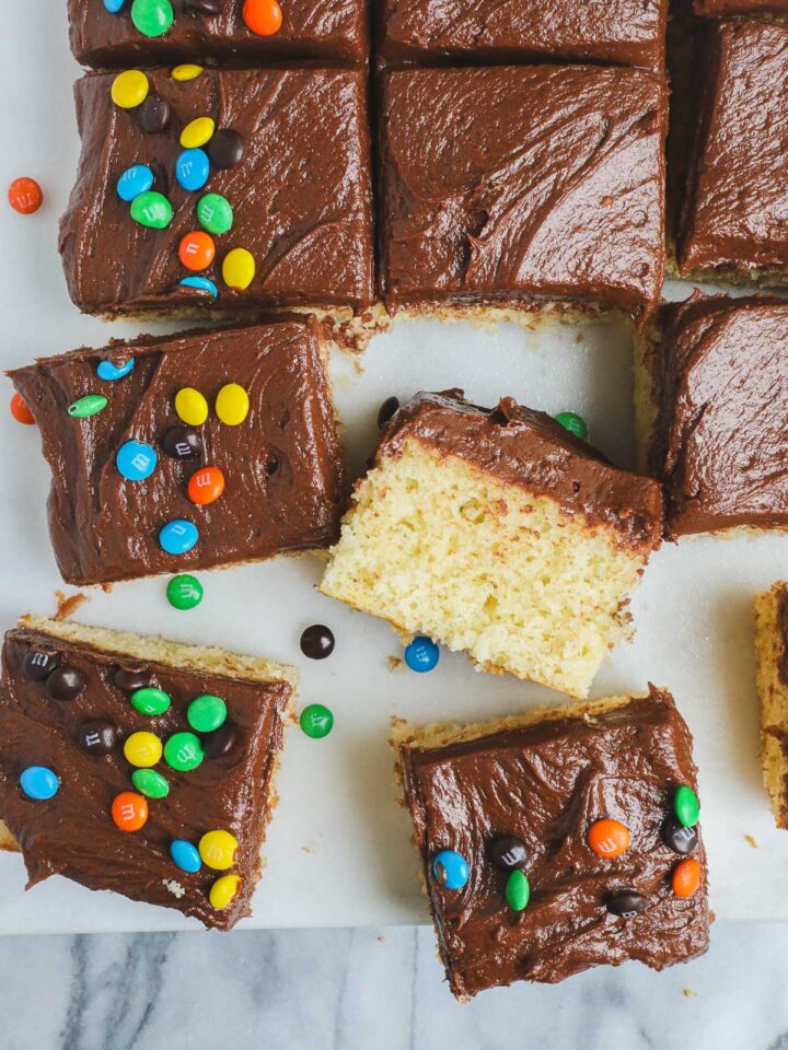 Cake Without A Mixer sliced into squares on a white marble serving board frosted with easy chocolate frosting and decorated with M&M's.