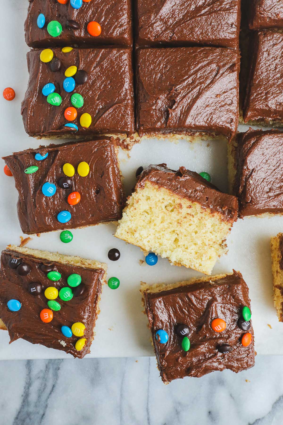 Cake Without A Mixer sliced into squares on a white marble serving board frosted with easy chocolate frosting and decorated with M&M's.