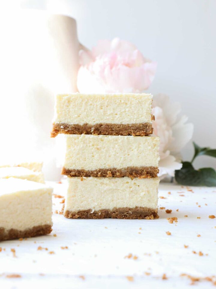 Three rectangular slices of cheesecake with a graham cracker crust stacked on top of each other with a light background.