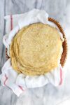 A large stack of twenty lightly browned einkorn tortillas layered on a towel in a bread basket.