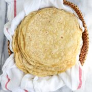 A large stack of twenty lightly browned einkorn tortillas layered on a towel in a bread basket.