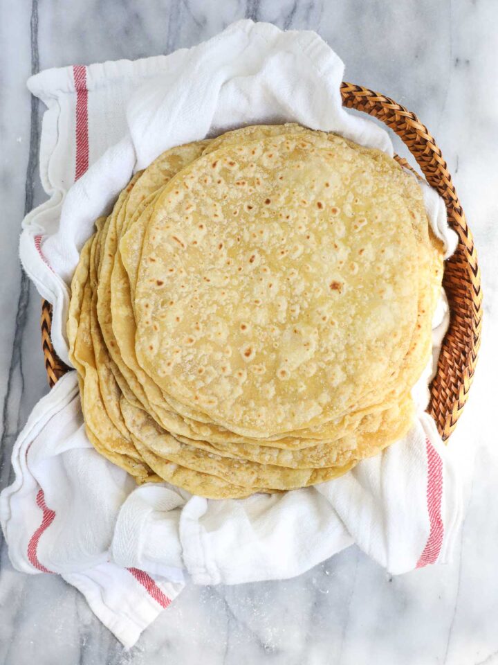 A large stack of twenty lightly browned einkorn tortillas layered on a towel in a bread basket.