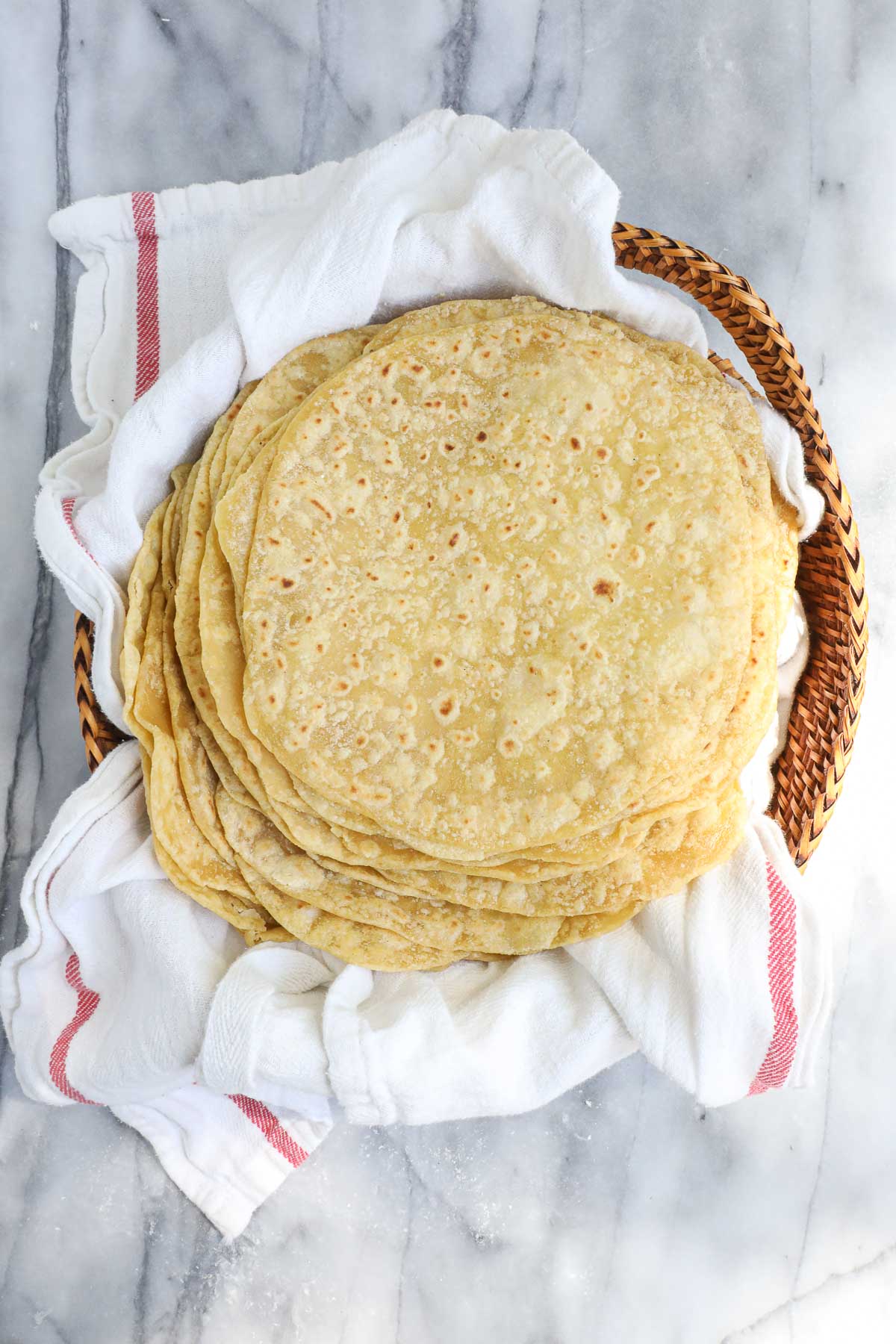 A large stack of twenty lightly browned einkorn tortillas layered on a towel in a bread basket.