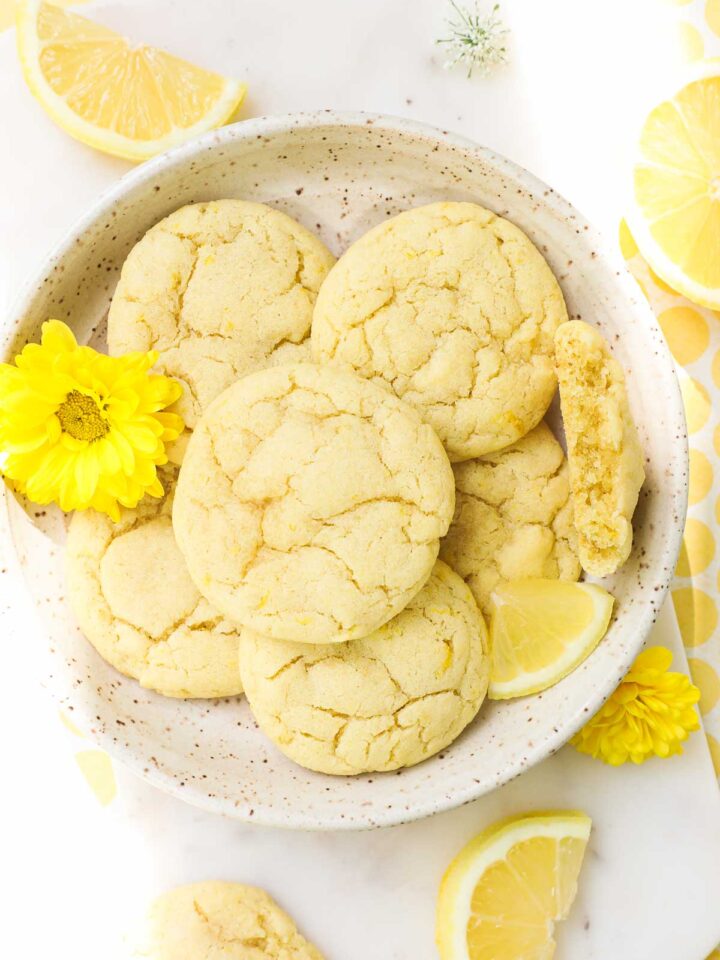 Lemon Olive Oil Cookies arranged on a plate on a white background decorated with yellow accents, lemons, and flowers.