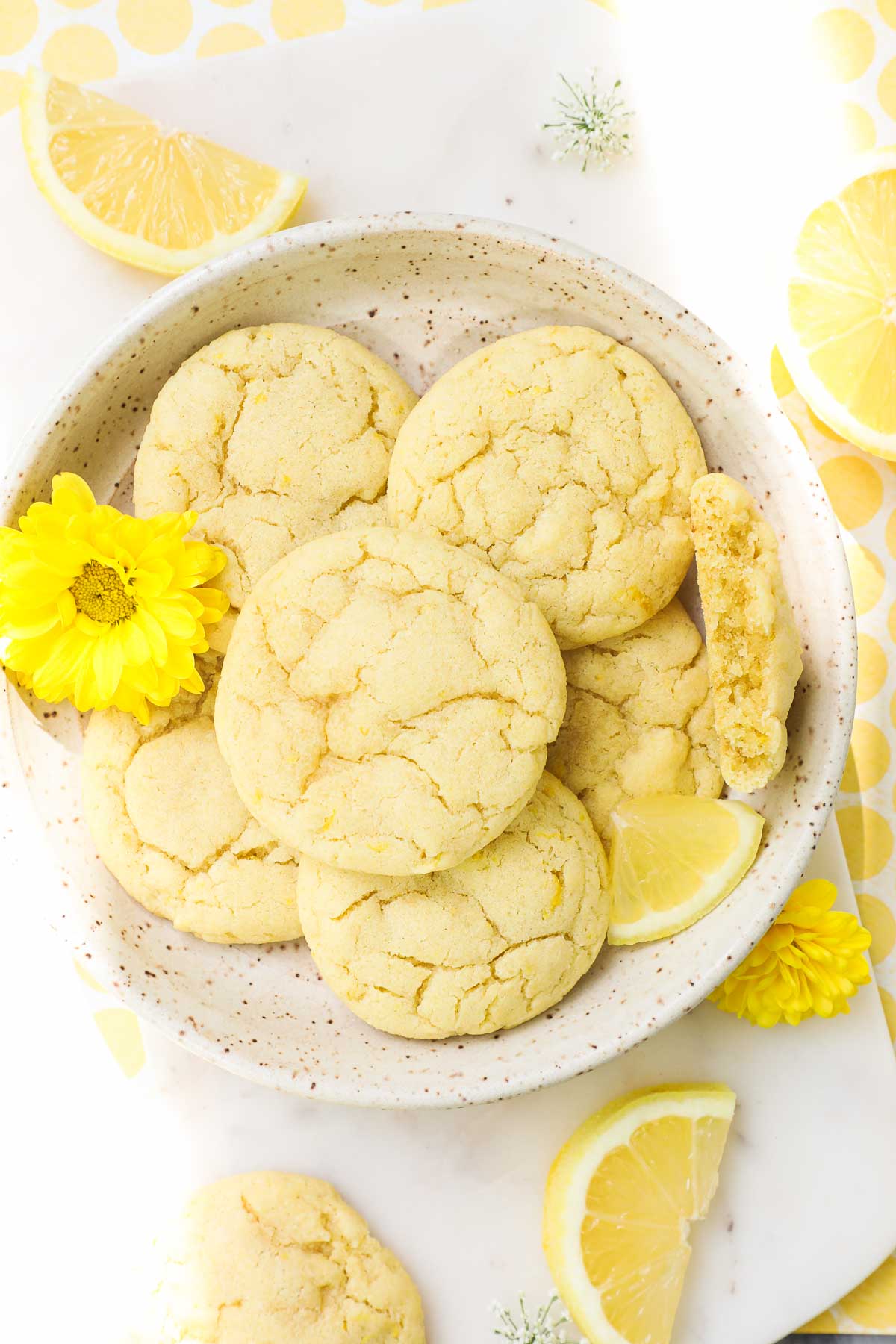 Lemon Olive Oil Cookies arranged on a plate on a white background decorated with yellow accents, lemons, and flowers.