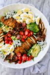 Crispy Chicken Cobb Salad arranged with groupings of sliced crispy chicken, sliced avocados, sliced hard boiled eggs, cherry tomatoes, bacon, and blue cheese on a bed of romaine lettuce.