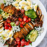Crispy Chicken Cobb Salad arranged with groupings of sliced crispy chicken, sliced avocados, sliced hard boiled eggs, cherry tomatoes, bacon, and blue cheese on a bed of romaine lettuce.