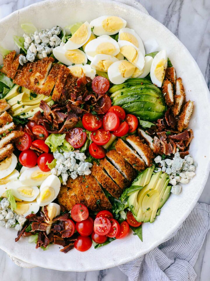 Crispy Chicken Cobb Salad arranged with groupings of sliced crispy chicken, sliced avocados, sliced hard boiled eggs, cherry tomatoes, bacon, and blue cheese on a bed of romaine lettuce.