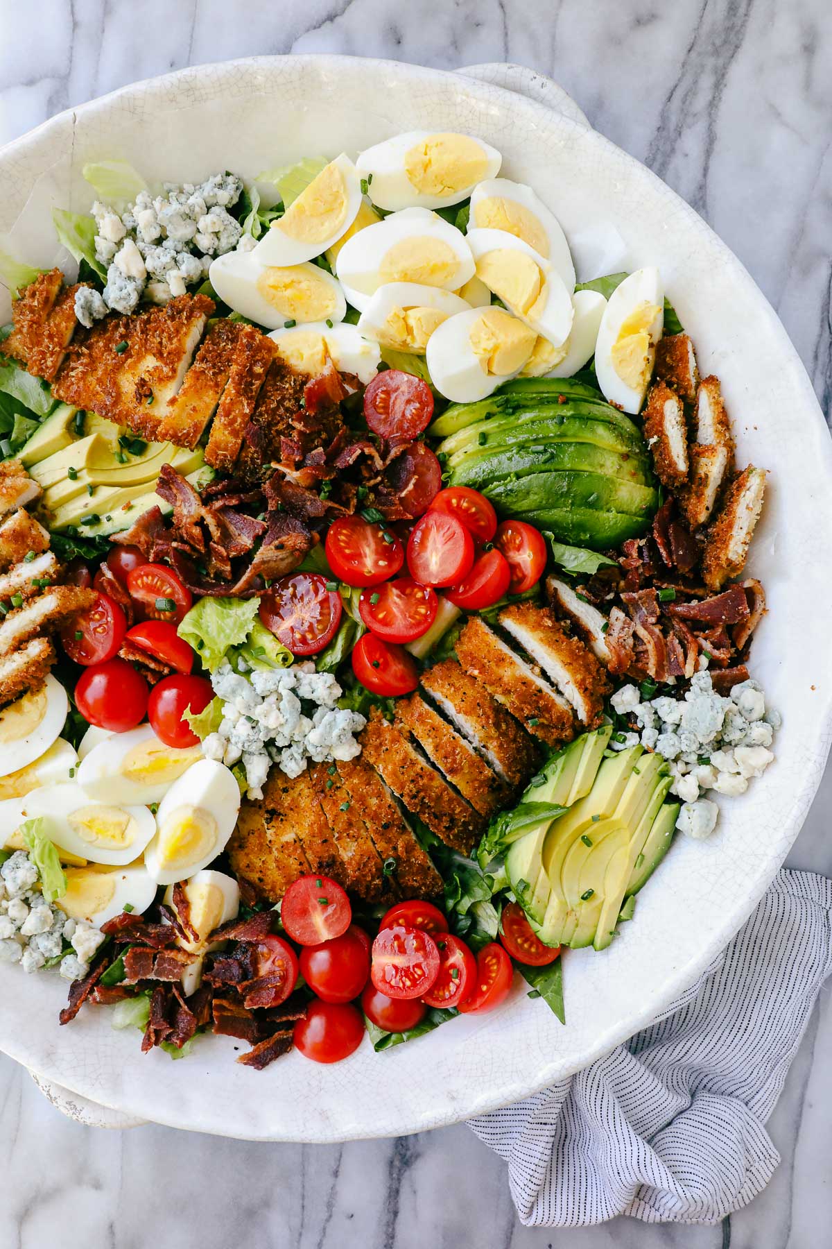 Crispy Chicken Cobb Salad arranged with groupings of sliced crispy chicken, sliced avocados, sliced hard boiled eggs, cherry tomatoes, bacon, and blue cheese on a bed of romaine lettuce.