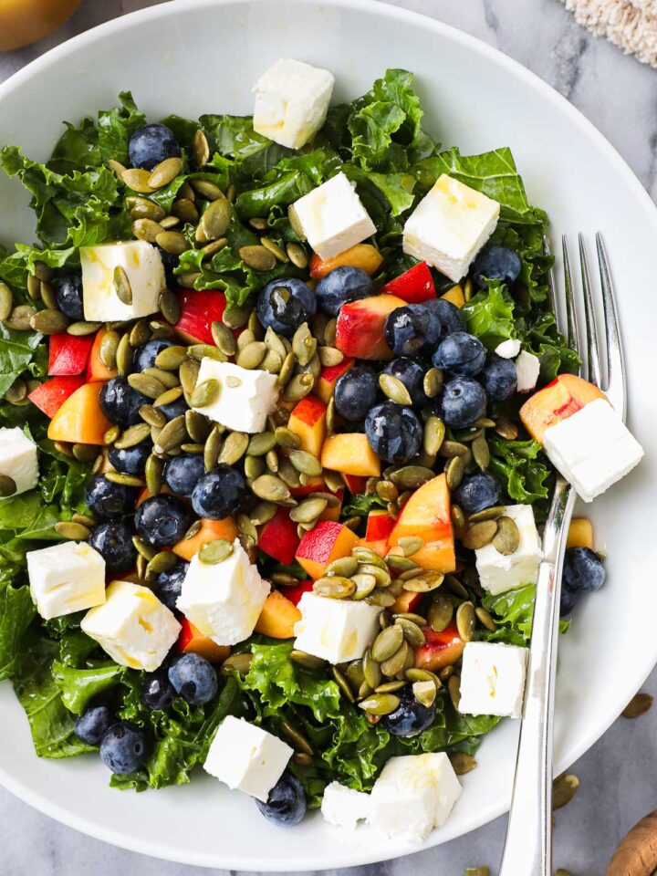 Summer Kale Salad in a bowl with diced nectarines, blueberries, feta, and pumpkin seeds with a tangerine vinaigrette overhead.