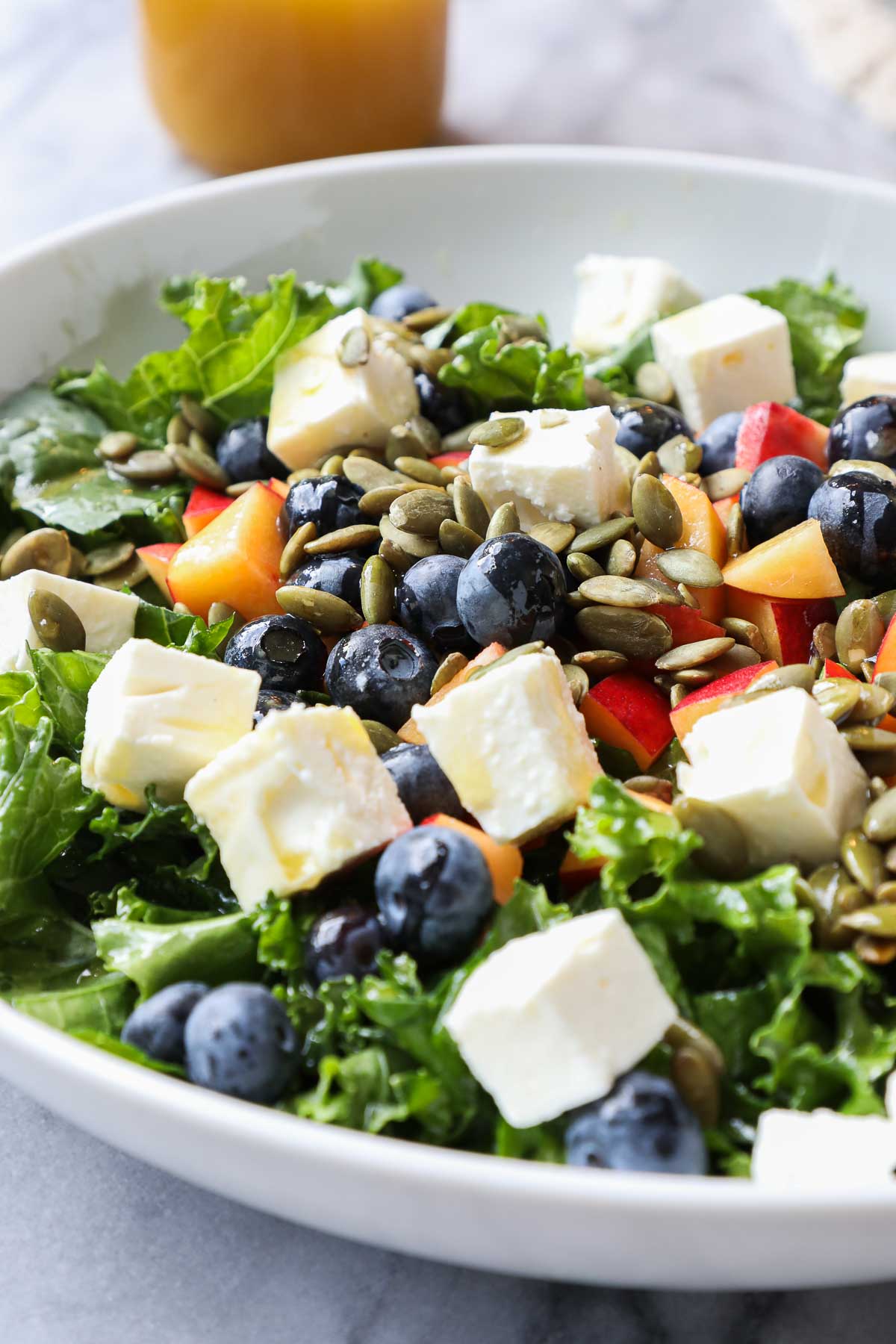 Summer Kale Salad in a bowl with diced nectarines, blueberries, feta, and pumpkin seeds with a tangerine vinaigrette at an angle.