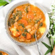 Tuscan White Bean Soup that is vibrant orange in color with cannellini beans, orange carrots, and green kale in a bowl on a soft cream colored napkin.
