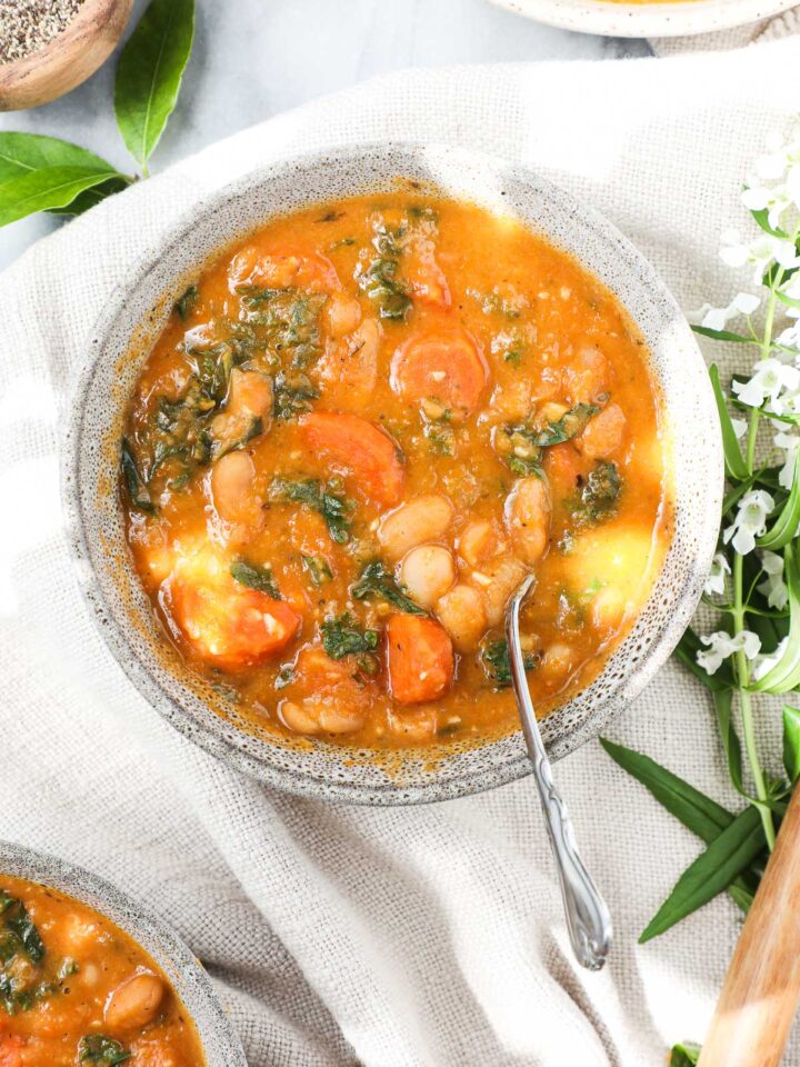 Tuscan White Bean Soup that is vibrant orange in color with cannellini beans, orange carrots, and green kale in a bowl on a soft cream colored napkin.