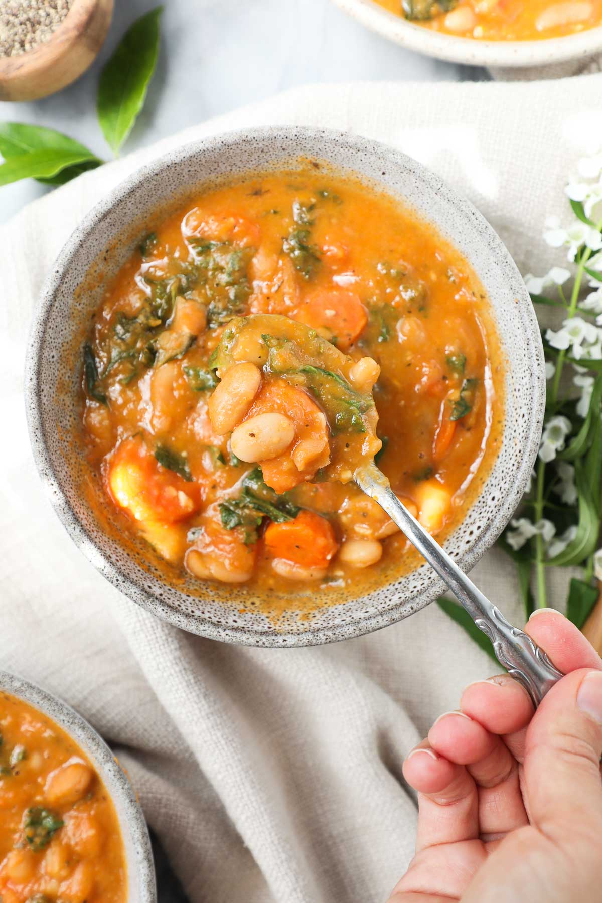 Lauren holding a spoonful of the Tuscan White Bean Soup over a bowl of soup on a light background styled with additional bowls of soup and greenery.