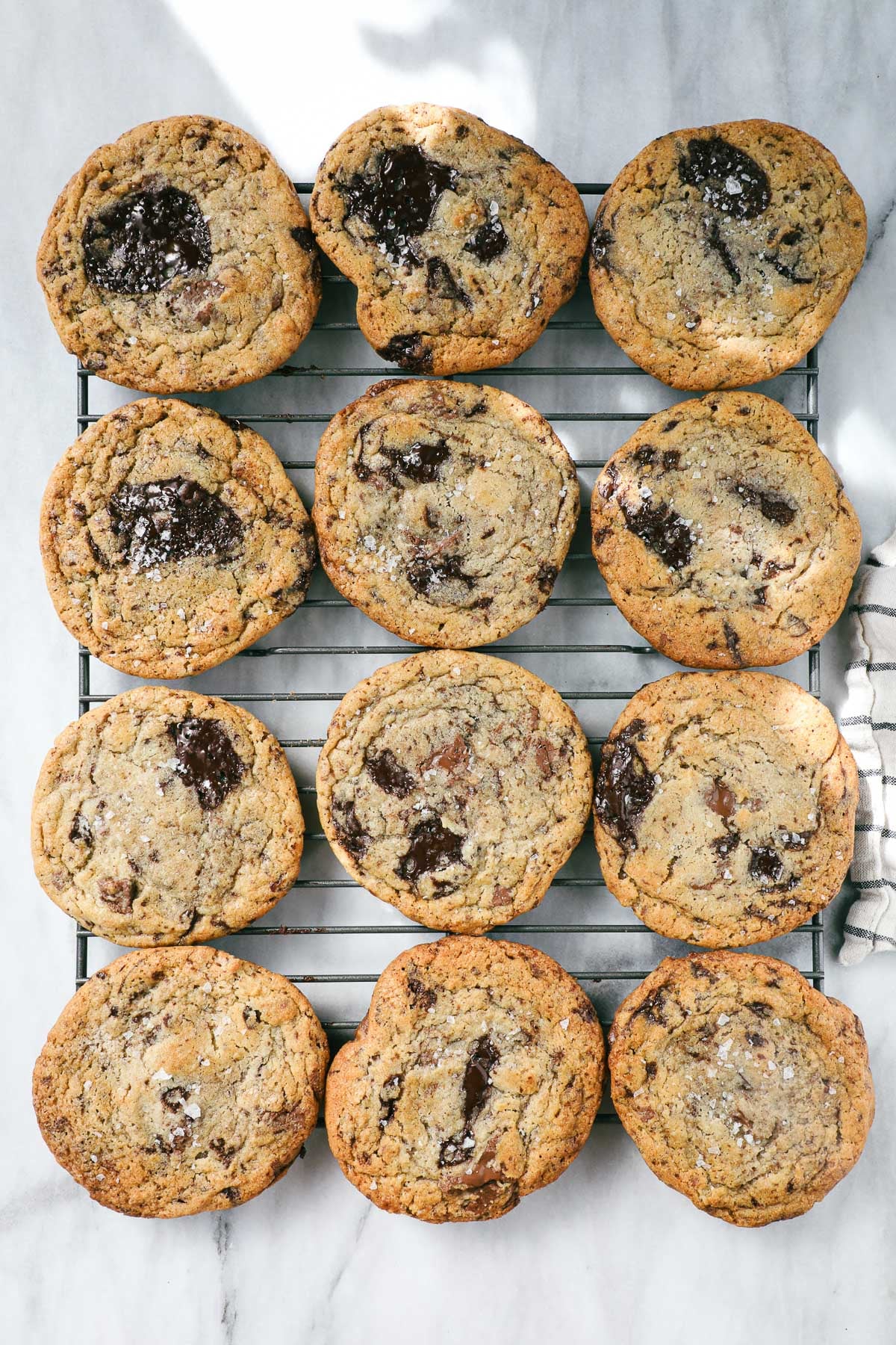 A dozen large chocolate chip cookies with melted pools of chocolate on a cookie drying rack on a white marble background.