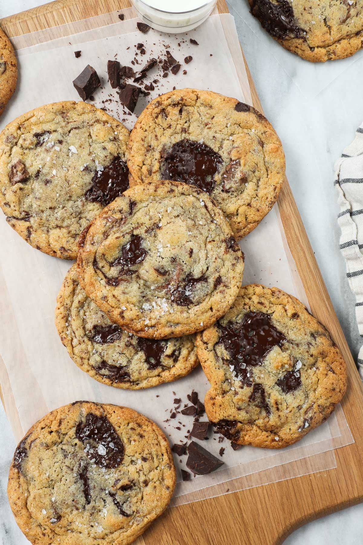 Large chocolate chip cookie arranged on a wooden serving board styled with chunks of chocolate and a white and grey striped napkin.