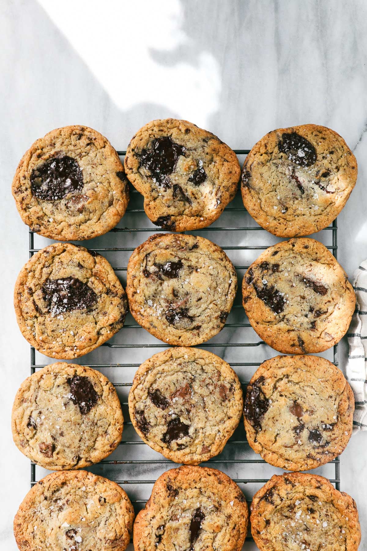 Twelve Cookies arranged in four rows of three on a rectangular cookie drying rack on a white marble surface dappled with natural light.