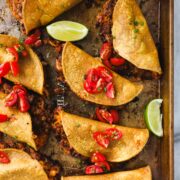 A golden baking sheet arranged with Crispy Oven Baked Chili Tacos with Ground beef layered with diced tomatoes and styled with sliced limes and guacamole.