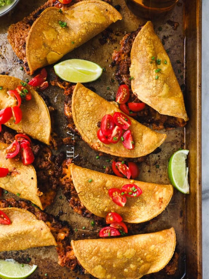 A golden baking sheet arranged with Crispy Oven Baked Chili Tacos with Ground beef layered with diced tomatoes and styled with sliced limes and guacamole.