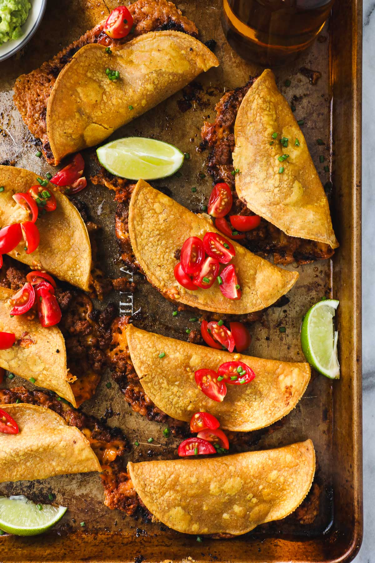 A golden baking sheet arranged with Crispy Oven Baked Chili Tacos with Ground beef layered with diced tomatoes and styled with sliced limes and guacamole.
