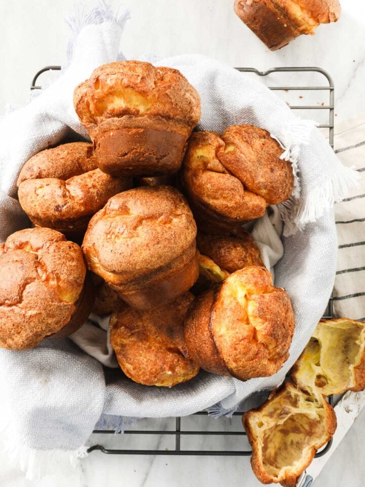 Popovers in a bread basket lined with a soft linen napkin on top of a drying rack on a white marble background.
