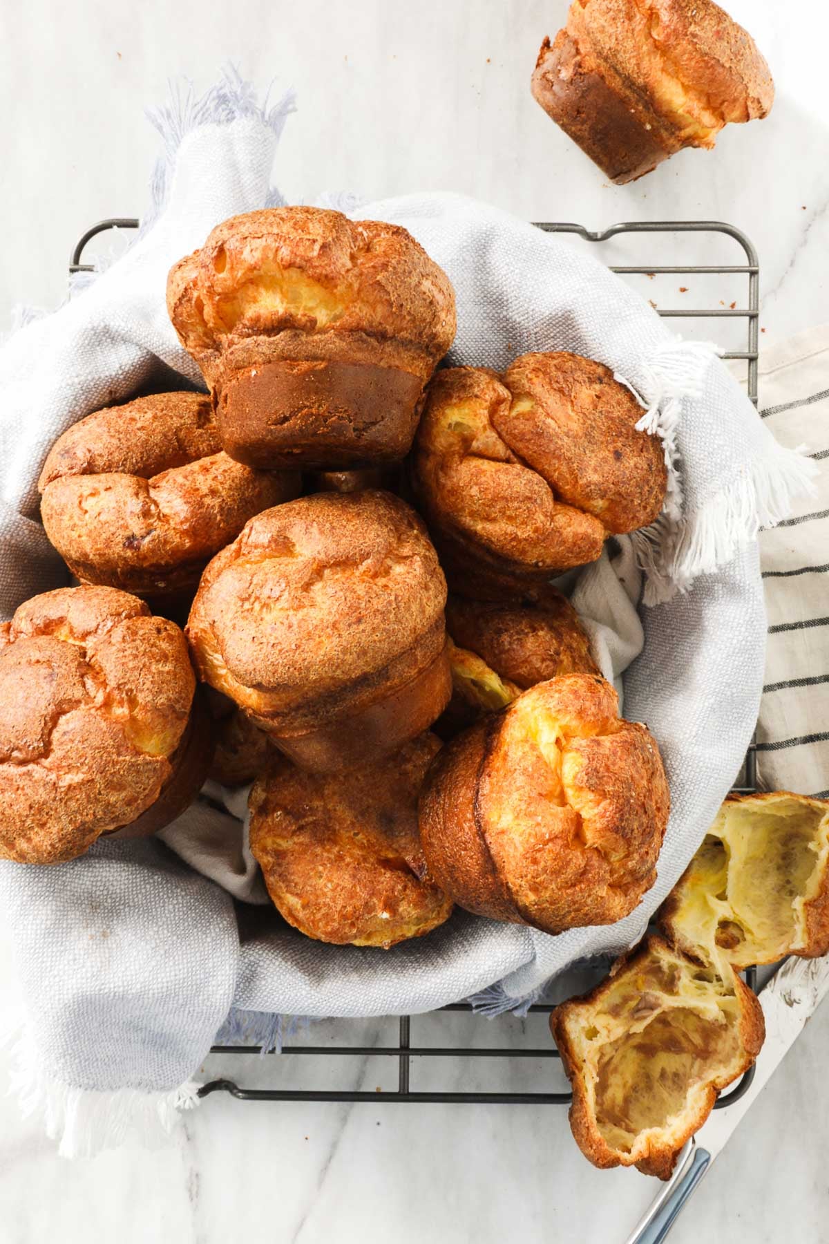 Popovers in a bread basket lined with a soft linen napkin on top of a drying rack on a white marble background.