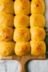 Golden Pumpkin Dinner Rolls in five rows of three on wooden cutting board on a marble background.