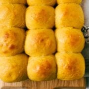 Golden Pumpkin Dinner Rolls in five rows of three on wooden cutting board on a marble background.