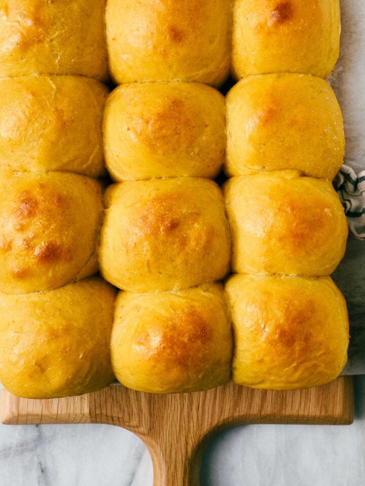 Golden Pumpkin Dinner Rolls in five rows of three on wooden cutting board on a marble background.