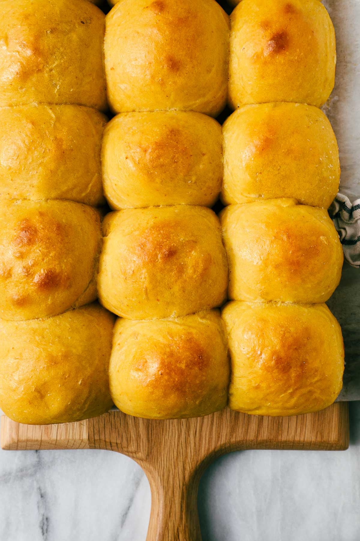 Golden Pumpkin Dinner Rolls in five rows of three on wooden cutting board on a marble background.
